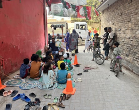 A cycle cart is seen at the end of an alley, with children seated in front of it and a few adults leaning against walls listening