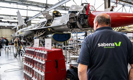 A man wearing a T-shirt with the logo ‘Sabena engineering’ stands with his back to the camera. He is in a workshop with part of an F-16 in front of him