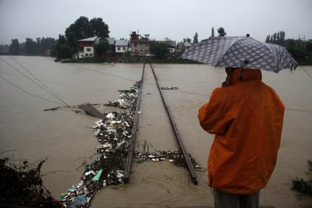 A person ion n orange jacket and sheltering under spotted umbrella looks out across a river that has overflowed its banks