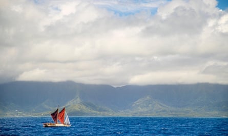 A two-masted boat in full sail passing a coastline of green hills topped by clouds