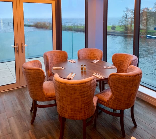 A dining table next to floor to ceiling windows overlooks a large lake.