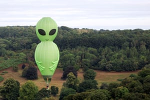 An alien-shaped green hot air balloon with black eyes stands in a field with trees.