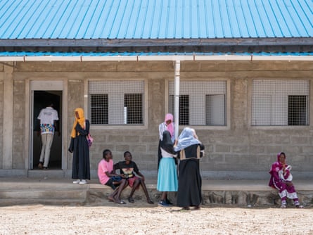 Children sit and stand outside a brick building with a corrugated roof. The girls wear scarves over their heads.