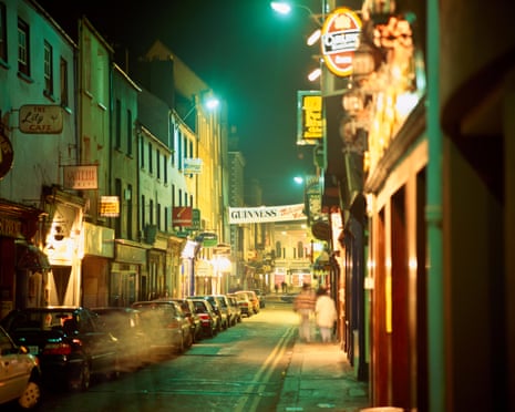 A street in Cork at night, illuminated by pub signs and street lights, with blurred images of people walking the streets and crossing the road. A Guinness banner hangs across the street in the distance.