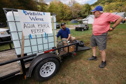 Man sits on tailgate of trailer alongside huge water carton with sign that says Drinking Water/Agua Para Beber, as a stand stands beside it, the two of them talking.