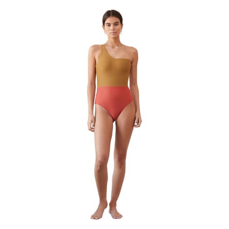 A model wears a Casa Raki Maillot one-shoulder swimsuit. The top half above the seam is in ochre and the bottom half is in coral.
