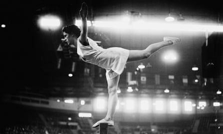 Black and white photo: a young woman balances on one leg on a bar, body horizontal, left leg stretched out behind her, arms outstretched. She wears a white singlet top and shorts, and thin slipper shoes.