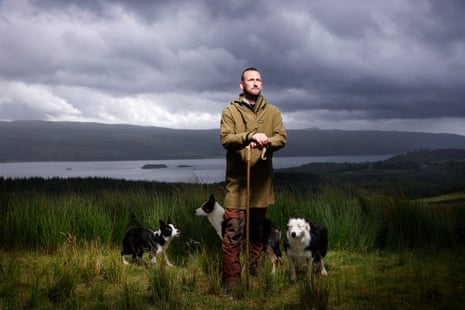 A man leans on a shepherd's crook with three dogs at his feet and a body of water in the background.
