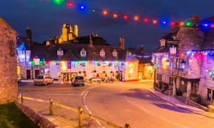 View of the lit-up ruin of Corfe Castle in Dorset, UK with Christmas lights decorating the village below.below.