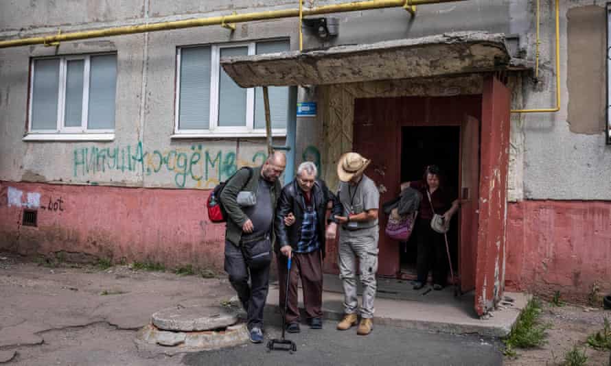 A man with a cane is helped out the door of a block of flats by two men, one wearing a cowboy hat
