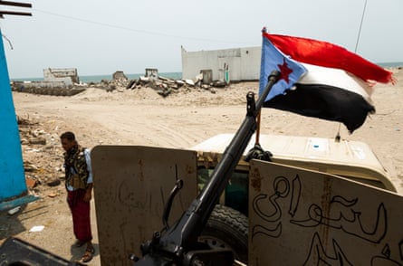 A man with ammunition pouches standing by a truck with a heavy machine-gun mounted on the back. Ruined buildings are in the background