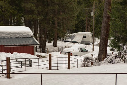 A house, car and RV can be seen covered in snow in an area surrounded by trees.