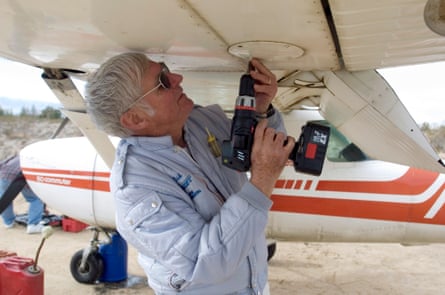An older man operates a battery-powered, handheld drill beneath the white wing of a biplane.