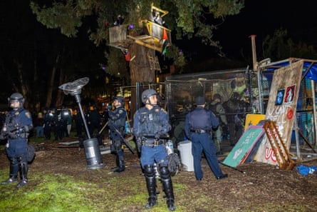 Lit perhaps by the flash of the camera, police officers in helmets stand around a tree near brightly painted plywood.
