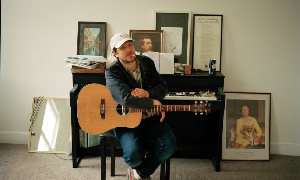Jamie T with guitar on lap and cigarette in mouth, in front of a piano dotted with portraits