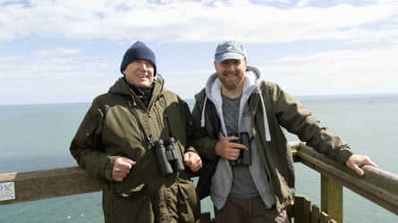 A man with a dark green winter jacket and grey woolly hat and a man with a green hooded jacket and pale blue cap both holding binoculars lean on a fence smiling
