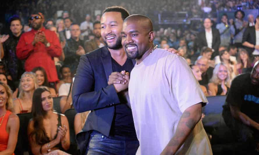 Two musicians, John Legend and Kanye West, shake hands as they perform in a crowded place.