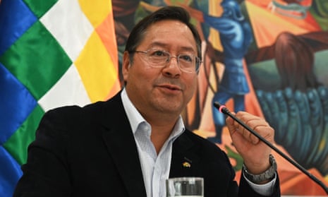 A middle-aged Bolivian man with trim black hair, a dark blazer and shirt open at the collar without a tie sits at a small microphone in front of a brightly colored flag of blue, green, white, yellow and orange.