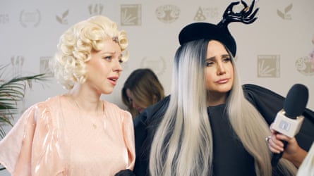 Ellie White and Natasia Demetriou in elaborate wigs and costumes, holding a microphone