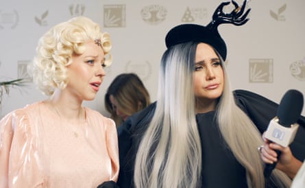 A woman in peach dress with short blond wig and a woman in black dress and fascinator look at an interviewer holding a mic up to them.