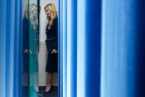 Press secretary Katie Zacharia walks through blue curtains holding a folder, reflected in glass