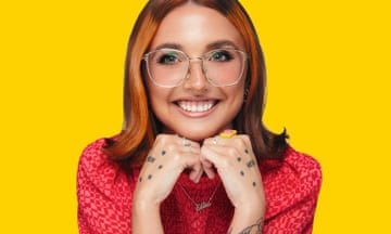 Ellie is white and has shoulder-length auburn hair in a centre parting, large glasses and a big smile. She is resting her chin on her hands which have tattoos and rings