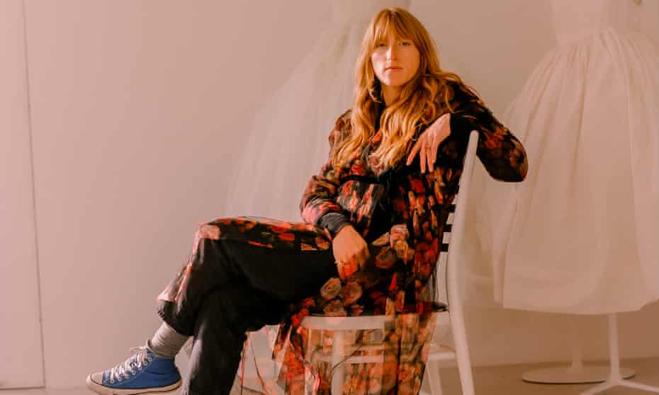 Molly Goddard, sitting, wearing black trousers and a black top with a long, colourful, fine fabric, transparent shirtdress over the top and blue lace-up canvas boots