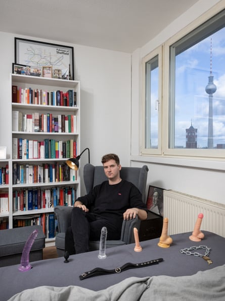 Portrait of Daniel sitting in a chair in the corner of a room with a bookcase and window behind him and a row of sex toys laid out on a table in front of him