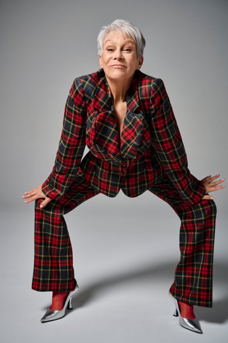 Portrait of Jamie Lee Curtis in a tartan suit and silver shoes crouching down with her hands on her knees