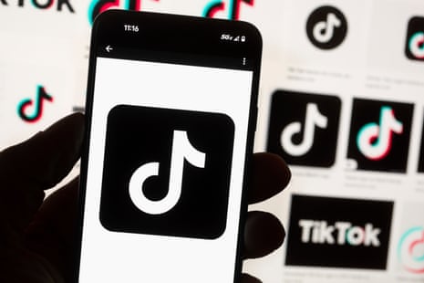 The TikTok logo is seen on a mobile phone in front of a computer screen which displays the TikTok home screen.