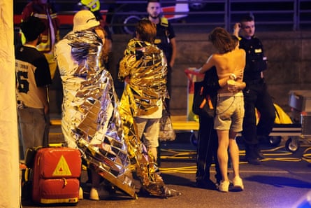 Two people embrace near others wearing thermal blankets as they stand near emergency personnel