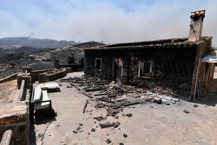 A house destroyed by the fire in Los Gallardos. It is a single-storey long, narrow building with its roof burned through and stone walls blackened. Debris is strewn on a terrace in front. Smoke rises from the hills behind.