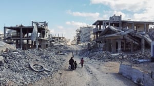 People walk along a dust road between piles of rubble and the bombed-out shells of buildings