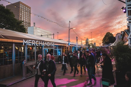 Crowds walking past a merchandise stall and bars at sunset.