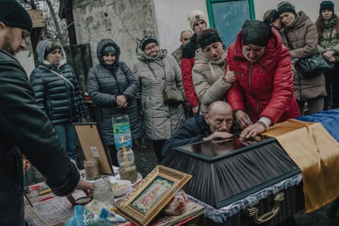 An elderly man cries as he kneels at the side of the coffin, while two women stand alongside, one with her hand on the coffin. Other mourners stand behind
