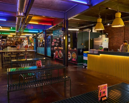 A low lit food hall with neon lighs and several stalls with rows of shared tables.