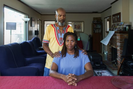 Woman sits with fingers interlaced on red table, as man stands behind her with hands on her shoulders