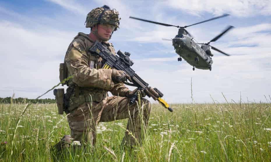 British soldier kneeling in field with helicopter taking off in background.