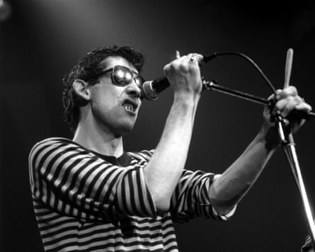 Shane MacGowan grimaces on stage while holding a stand microphone and wearing dark sunglasses