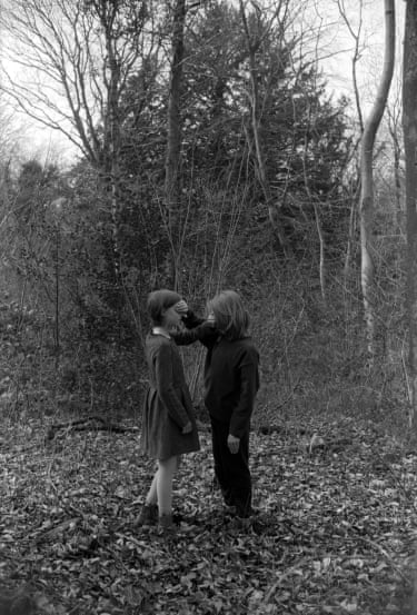 Black and white photograph of two children standing in the middle of a forest. The person on the right is obscuring the eyes of the person on the left, and the person on the left is obscuring the eyes of the person on the right