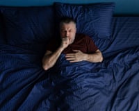 A middle aged man coughing while lying in bed under a blue blanket.