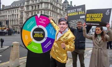 A person dressed as Rishi Sunak as a gameshow host with the 'wheel of misfortune' as people hold signs protesting against the Rwanda bill