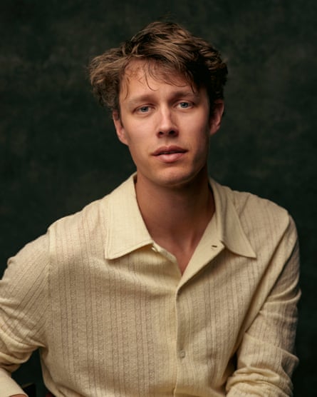 he is in his mid-30s with light brown hair which is long on top and falls across his forehead. He wears a pale yellow open-necked shirt