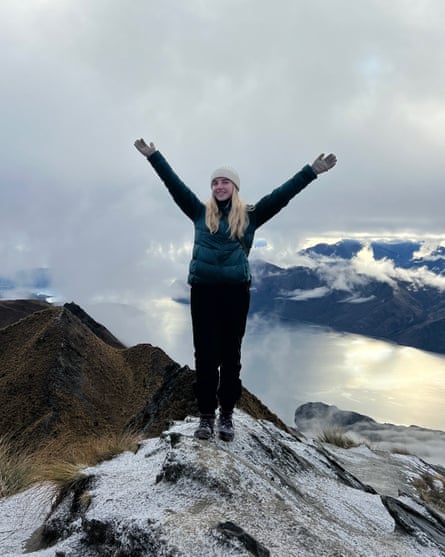 Wearing winter gear, she stands in a Y-shape on a snow-topped peak with mountains and a large lake in the background