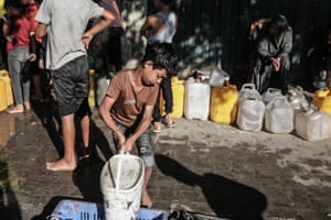 A small boy picks up a large white container as people stand or sit near large water containers