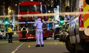 A double decker bus beside a police officer in a hi-viz jacket, with a police vehicle to the right and an officer in a hazmat suit centre foreground. Lines of police tape cordon off the apparent night time crime scene.