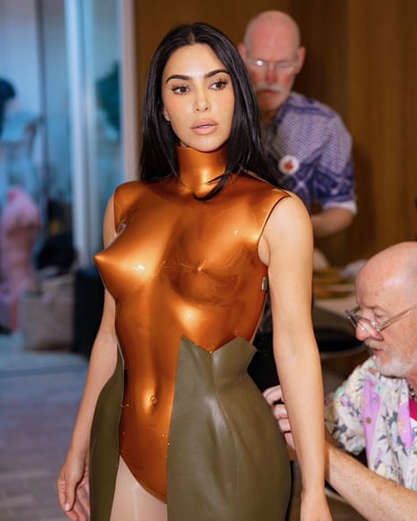 Kim Kardashian in a metallic bronze sculptural bodysuit during a fitting with two male designers behind her.