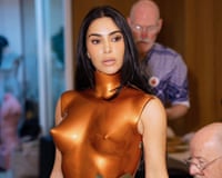 Kim Kardashian in a metallic bronze sculptural bodysuit during a fitting with two male designers behind her.