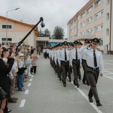 Graduating cadets march in formation as parents and family members watch. They are in a courtyard between cream and pale orange painted buildings.