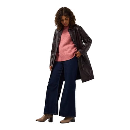 A model wears a Brown Patent Faux Leather Coat with jeans, boots and a pink jumper. They have shoulder length brown hair and are looking down.